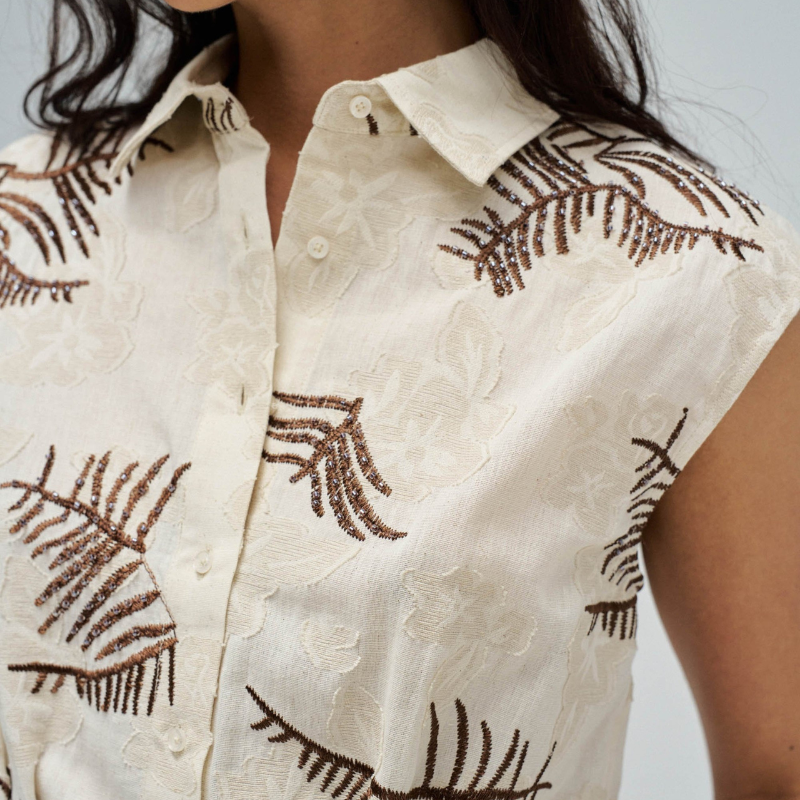 Camisa sem mangas em tons bege e castanho com detalhes sofisticados de bordados e missangas. Corte slim que segue a silhueta, com decote em colarinho clássico e abertura frontal com botões funcionais. Pregas estratégicas na cintura adicionam movimento e conforto. Peça versátil perfeita para looks casuais ou mais elaborados.