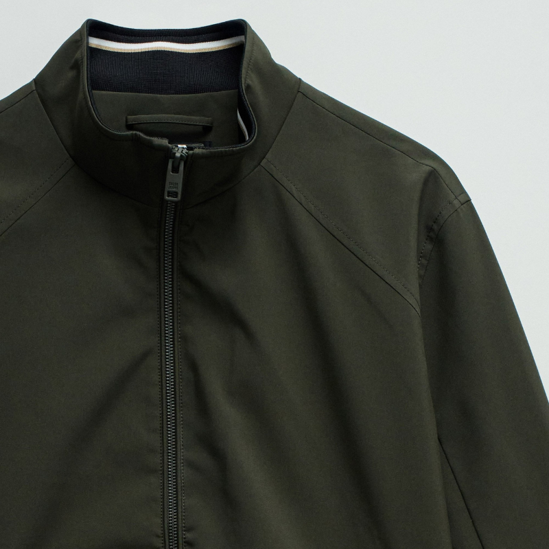 Casaco bomber em verde escuro com design versátil e funcional. Apresenta abertura frontal com fecho de correr para ajuste fácil, fit regular que se adapta confortavelmente ao corpo, e mangas compridas com punhos elásticos que garantem um acabamento limpo. Equipado com dois bolsos laterais práticos e bolso interior com fecho para maior segurança dos seus pertences. O elástico na cintura proporciona um corte definido e moderno. Uma peça essencial do guarda-roupa que combina estilo casual com funcionalidade.