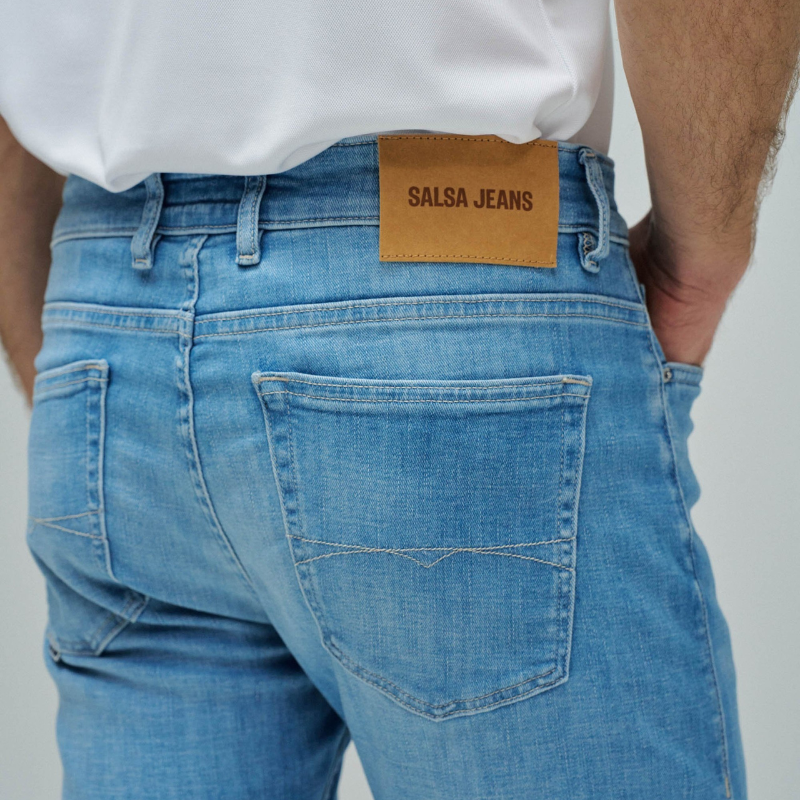 Calças jeans slim fit em azul claro com cintura média e perna ajustada. Apresenta abertura com botões, cinco bolsos funcionais e presilhas para cinto. Corte moderno que realça a silhueta, ideal para um visual casual e versátil no dia a dia.