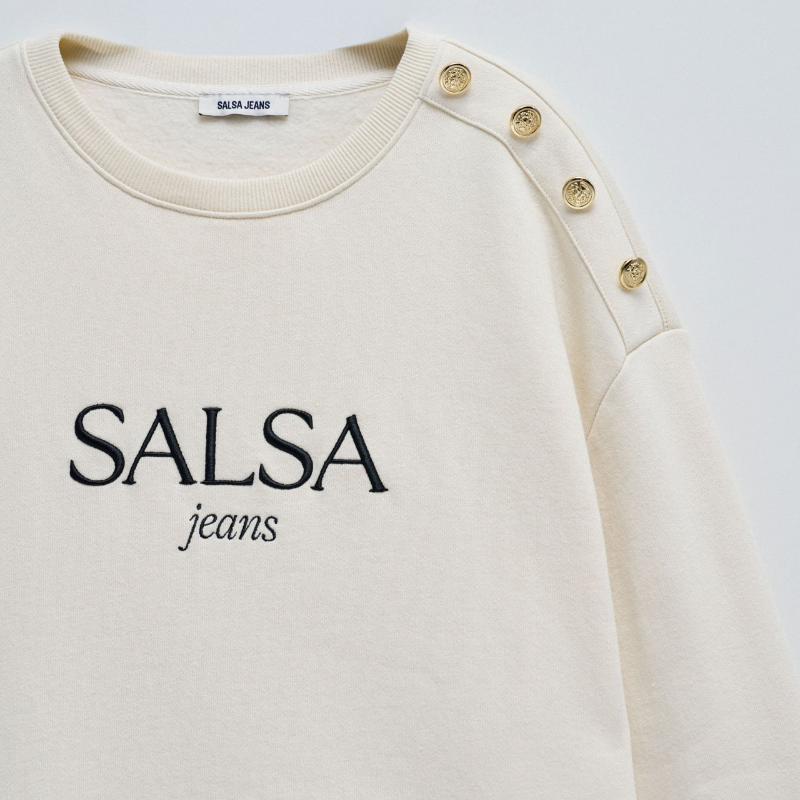 Camisola de felpa em bege com branding em preto. Camisola de mangas compridas com punhos canelados e decote redondo. Detalhe de quatro botões dourados num ombro.
Corte: Regular fit