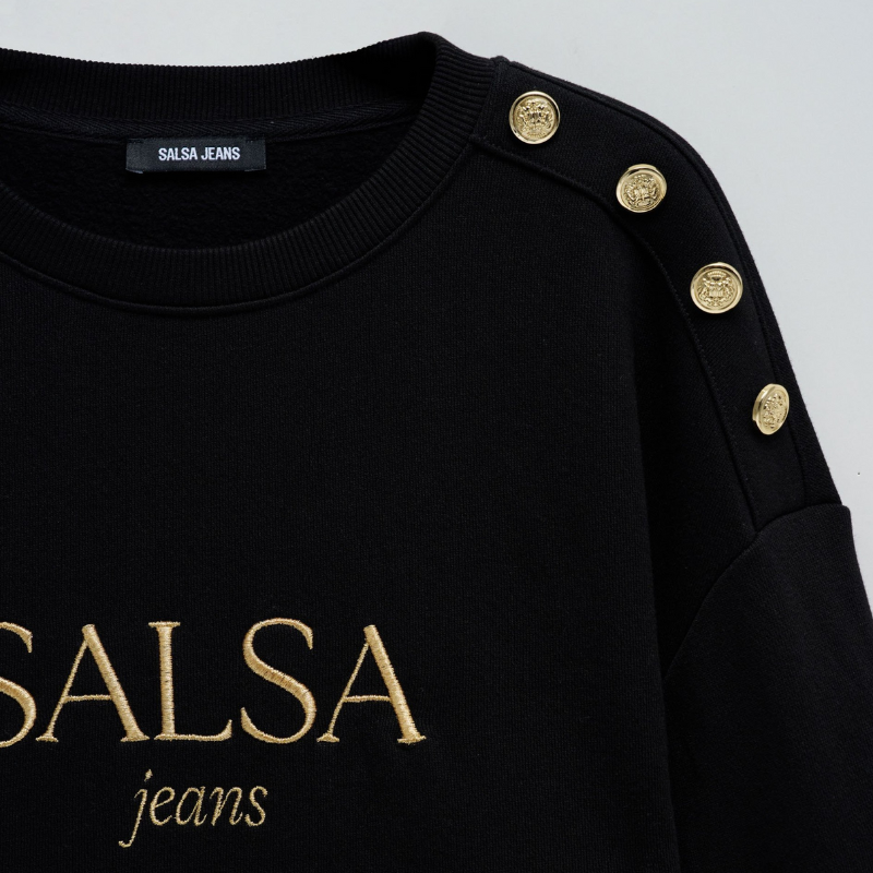 Camisola de felpa em preto com branding em dourado. Camisola de mangas compridas com punhos canelados e decote redondo. Detalhe de quatro botões dourados num ombro.
Corte: Regular fit