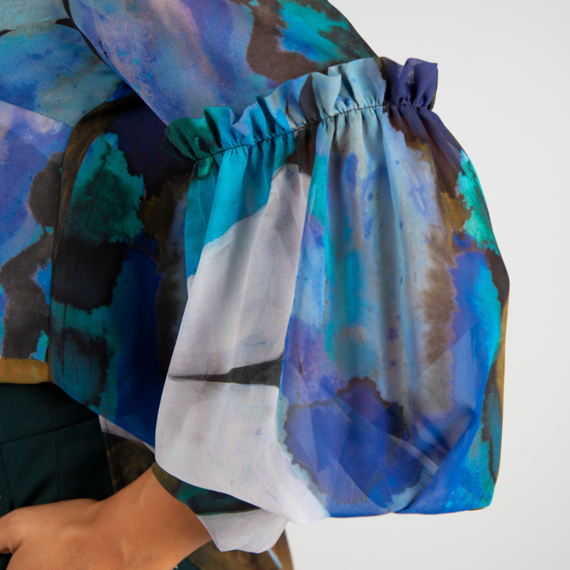 Túnica de decote em V com estampado abstrato em tons de turquesa, azul e verde. Túnica de mangas compridas em formato balão, levemente transparentes e forrada no corpo. Mangas a terminar com punho alto com dois botões metálicos em relevo.
Composição: 100%Poliester