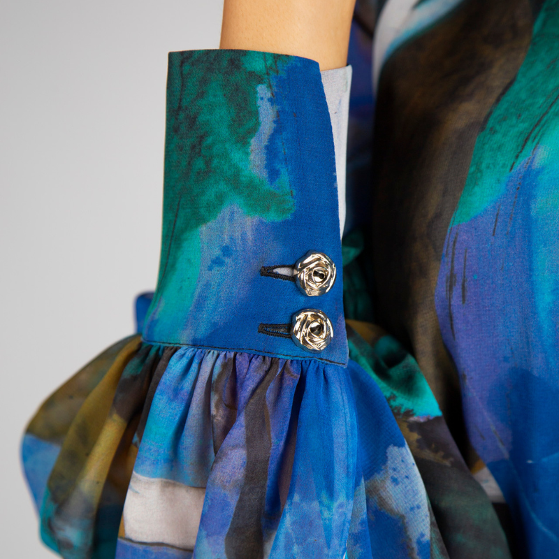 Túnica de decote em V com estampado abstrato em tons de turquesa, azul e verde. Túnica de mangas compridas em formato balão, levemente transparentes e forrada no corpo. Mangas a terminar com punho alto com dois botões metálicos em relevo.
Composição: 100%Poliester