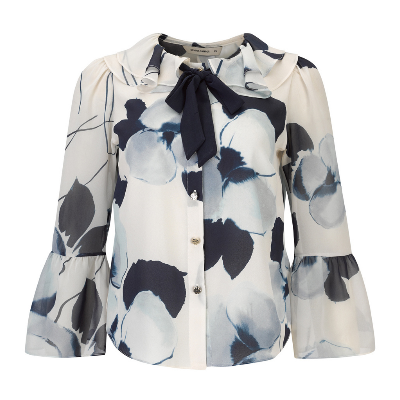 Blusa com estampado floral abstrato com padrão, em tons de azul-marinho, azul-claro e branco, confere um efeito delicado e atual ao visual. A gola com folhos traz um toque romântico, enquanto o laço azul escuro amovível no decote permite adaptar o look conforme a ocasião. Fecho frontal por botões prata e mangas compridas com punhos em folho que completam o design com requinte e feminilidade.