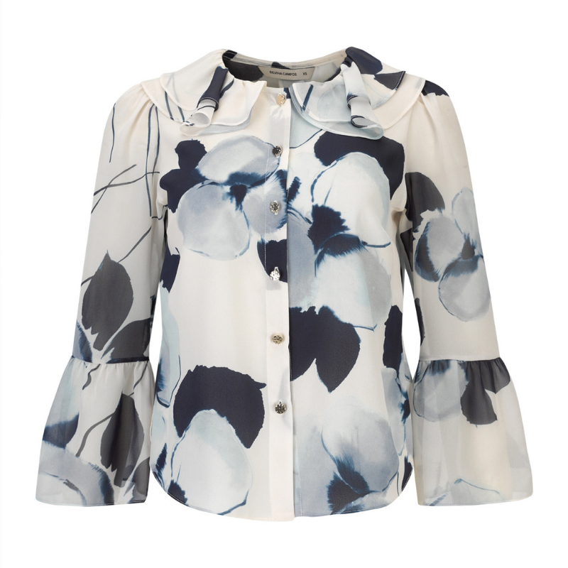 Blusa com estampado floral abstrato com padrão, em tons de azul-marinho, azul-claro e branco, confere um efeito delicado e atual ao visual. A gola com folhos traz um toque romântico, enquanto o laço azul escuro amovível no decote permite adaptar o look conforme a ocasião. Fecho frontal por botões prata e mangas compridas com punhos em folho que completam o design com requinte e feminilidade.