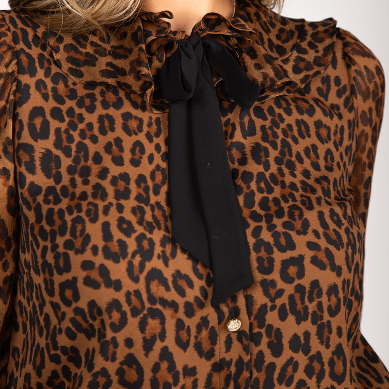Blusa com estampado animal print de leopardo em tons de castanho, caramelo e preto que conferem profundidade e sofisticação. Confecionada em tecido leve e semitransparente com caimento fluido e elegante. A gola com folhos traz um toque romântico, enquanto o laço preto amovível no decote permite adaptar o look conforme a ocasião. Fecho frontal por botões prata e mangas compridas com punhos em folho que completam o design com requinte e feminilidade.