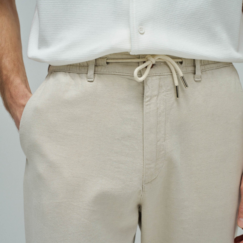 Calças joggers em algodão e linho que combinam conforto e estilo casual. O tecido natural em tom crú oferece leveza perfeita para dias quentes, enquanto a cintura média com cordão ajustável e elástico garante um encaixe seguro e confortável. Apresenta abertura com fecho de correr na cintura para facilitar o vestir, fit regular na perna e quatro bolsos funcionais. As presilhas completam o design prático e versátil, ideal para um look descontraído do dia a dia.