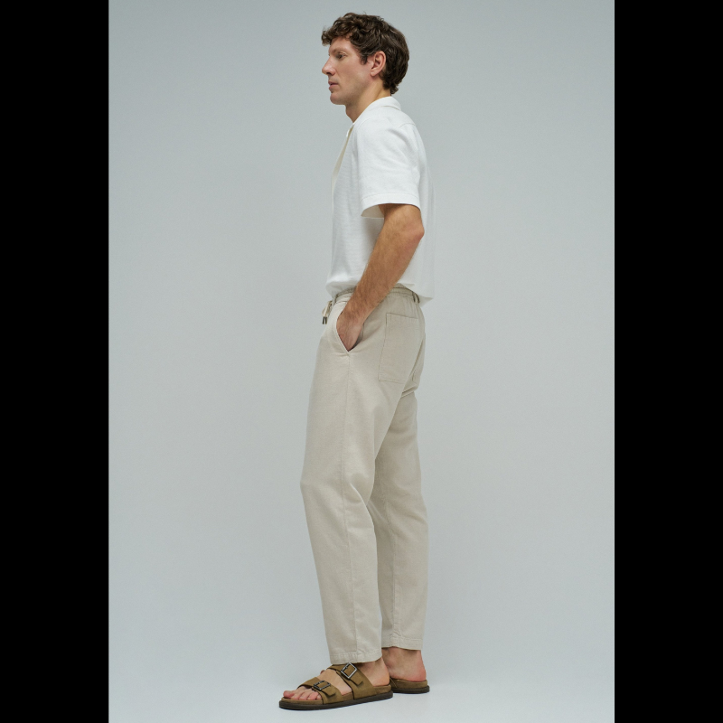Calças joggers em algodão e linho que combinam conforto e estilo casual. O tecido natural em tom crú oferece leveza perfeita para dias quentes, enquanto a cintura média com cordão ajustável e elástico garante um encaixe seguro e confortável. Apresenta abertura com fecho de correr na cintura para facilitar o vestir, fit regular na perna e quatro bolsos funcionais. As presilhas completam o design prático e versátil, ideal para um look descontraído do dia a dia.