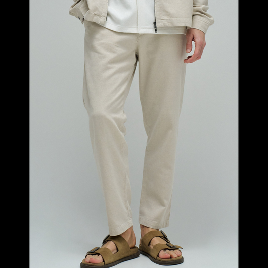 Calças joggers em algodão e linho que combinam conforto e estilo casual. O tecido natural em tom crú oferece leveza perfeita para dias quentes, enquanto a cintura média com cordão ajustável e elástico garante um encaixe seguro e confortável. Apresenta abertura com fecho de correr na cintura para facilitar o vestir, fit regular na perna e quatro bolsos funcionais. As presilhas completam o design prático e versátil, ideal para um look descontraído do dia a dia.