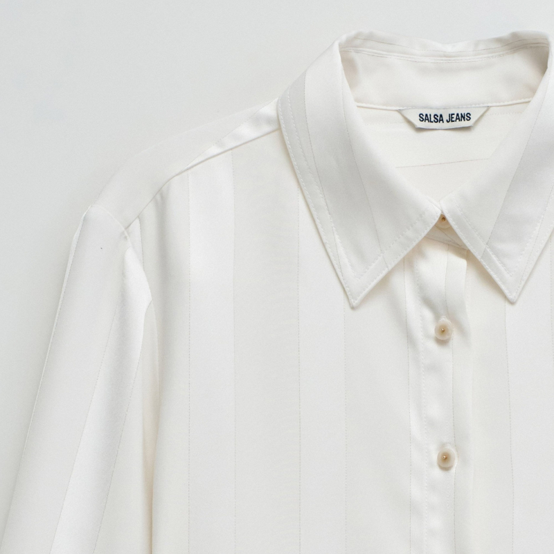 Camisa acetinada de corte regular com riscas em Lurex que adiciona um toque de sofisticação ao seu guarda-roupa. Confecionada em branco puro, apresenta mangas compridas e decote de colarinho clássico. O fecho frontal com botões e os punhos abotoados garantem um ajuste preciso e confortável. Ideal para ocasiões que exigem elegância discreta com um detalhe luminoso.