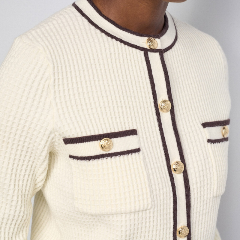 Cardigan versátil em tom bege com elegantes rebordos castanhos. Corte slim ajustado que valoriza a silhueta, mangas compridas e abertura frontal com botões dourados que adicionam um toque sofisticado. Os bolsos no peito reforçam a estética inspirada no tailoring francês. 