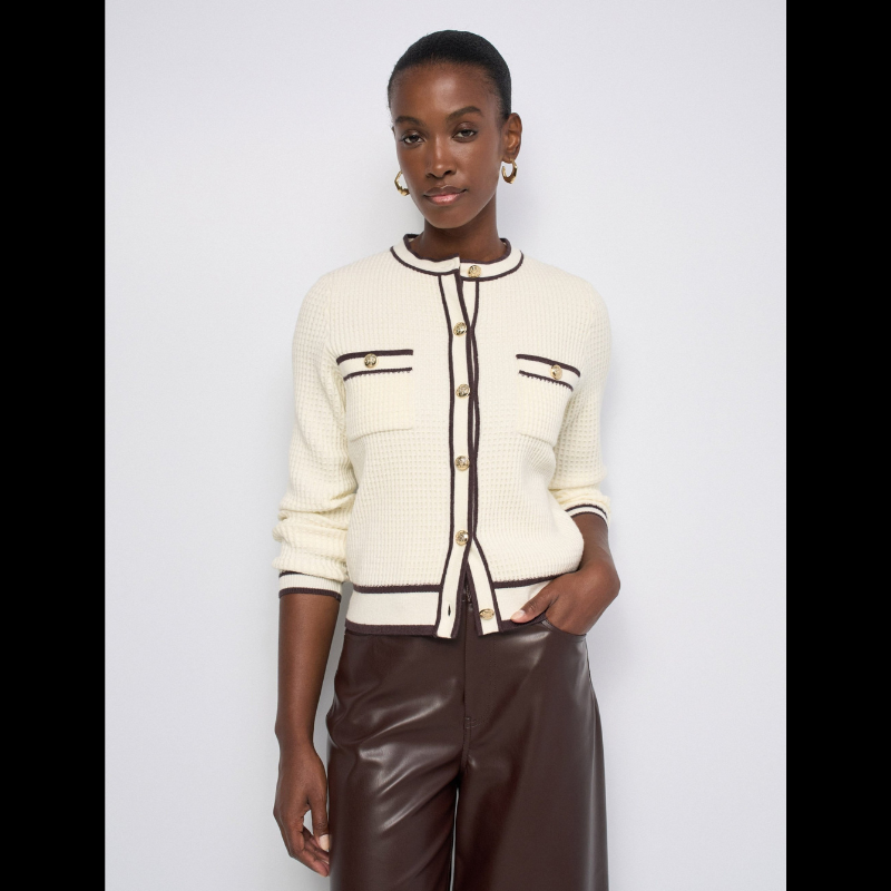 Cardigan versátil em tom bege com elegantes rebordos castanhos. Corte slim ajustado que valoriza a silhueta, mangas compridas e abertura frontal com botões dourados que adicionam um toque sofisticado. Os bolsos no peito reforçam a estética inspirada no tailoring francês. 