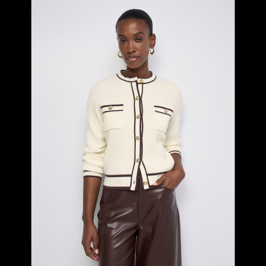 Cardigan versátil em tom bege com elegantes rebordos castanhos. Corte slim ajustado que valoriza a silhueta, mangas compridas e abertura frontal com botões dourados que adicionam um toque sofisticado. Os bolsos no peito reforçam a estética inspirada no tailoring francês. 