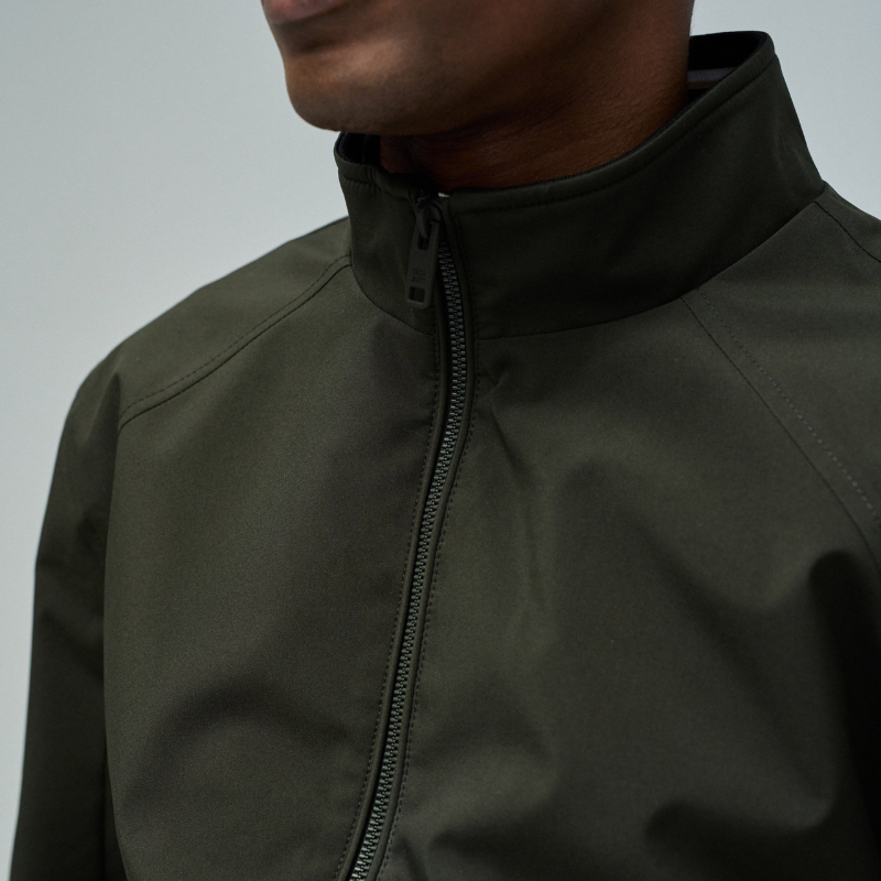 Casaco bomber em verde escuro com design versátil e funcional. Apresenta abertura frontal com fecho de correr para ajuste fácil, fit regular que se adapta confortavelmente ao corpo, e mangas compridas com punhos elásticos que garantem um acabamento limpo. Equipado com dois bolsos laterais práticos e bolso interior com fecho para maior segurança dos seus pertences. O elástico na cintura proporciona um corte definido e moderno. Uma peça essencial do guarda-roupa que combina estilo casual com funcionalidade.