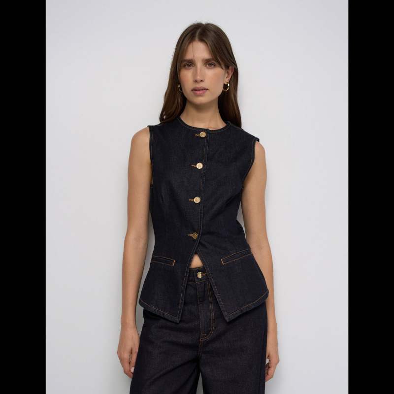 Colete em denim de cor azul escuro com um toque clássico e versátil. Corte de fit regular sem mangas, perfeito para criar camadas sobre as tuas peças favoritas. Destaca-se pela abertura frontal com botões dourados que adicionam um detalhe elegante e sofisticado. O decote redondo oferece conforto e fluidez, enquanto os dois bolsos não funcionais completam o design. 