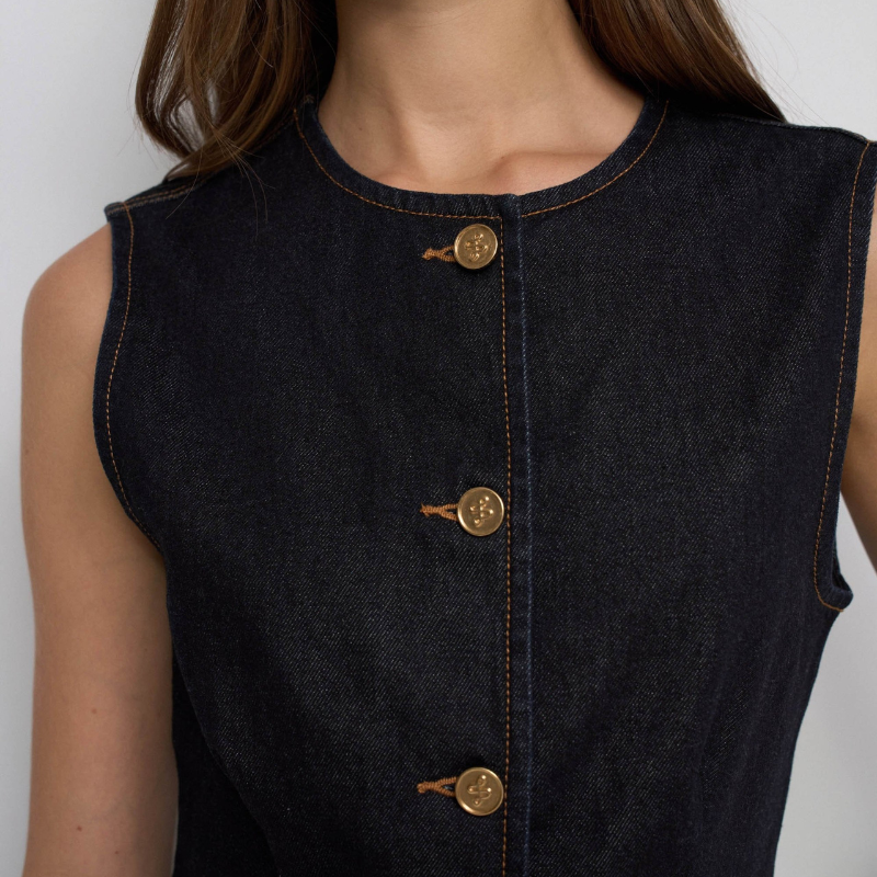 Colete em denim de cor azul escuro com um toque clássico e versátil. Corte de fit regular sem mangas, perfeito para criar camadas sobre as tuas peças favoritas. Destaca-se pela abertura frontal com botões dourados que adicionam um detalhe elegante e sofisticado. O decote redondo oferece conforto e fluidez, enquanto os dois bolsos não funcionais completam o design. 
