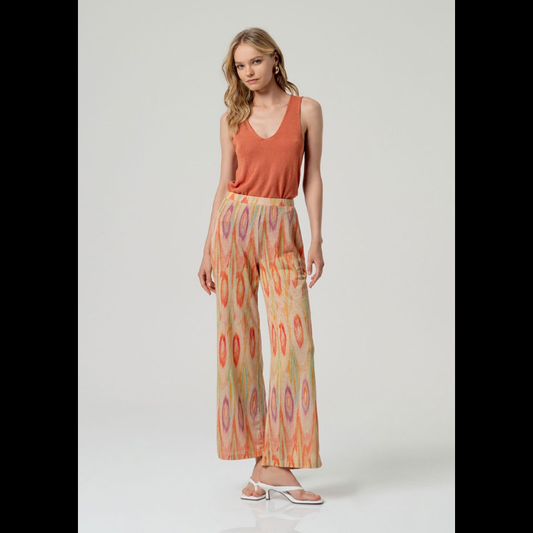 Elegantes calças pantalonas em malha fina da SURKANA, caracterizadas pela cintura média/alta com elástico na cintura que oferece conforto e ajuste seguro. O design apresenta um padrão geométrico em tons quentes de vermelho, laranja e verde sobre base bege, enriquecido com detalhes em Lurex dourado que conferem sofisticação e brilho. Peça versátil ideal para composições contemporâneas.