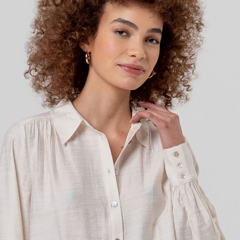 Camisa de manga comprida em crú fluida com gola e botões na frente. Ombros com franzidos que dão volume e punhos bufantes.
