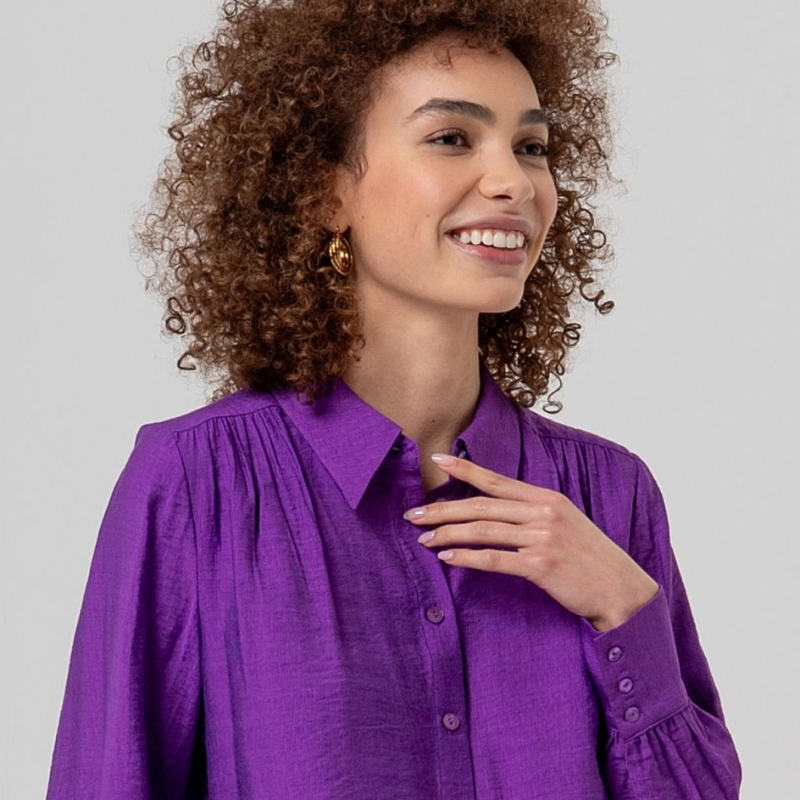 Camisa de manga comprida, em roxo, fluida com gola e botões na frente. Ombros com franzidos que dão volume e punhos bufantes.