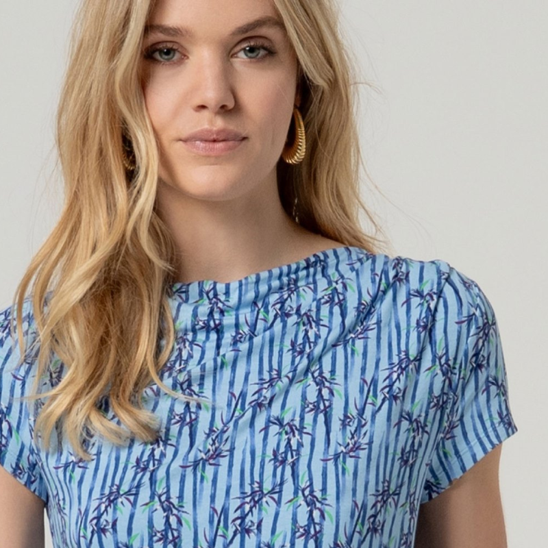 Camisola de manga curta em azul com decote a barco e drapeado elegante num ombro. Apresenta um padrão sofisticado que combina riscas verticais com motivos florais, criando um visual dinâmico e feminino. Confecionada em viscose EcoVero e elastano, oferece conforto e flexibilidade, com a vantagem de ser uma escolha sustentável. Perfeita para looks casuais ou mais elaborados.