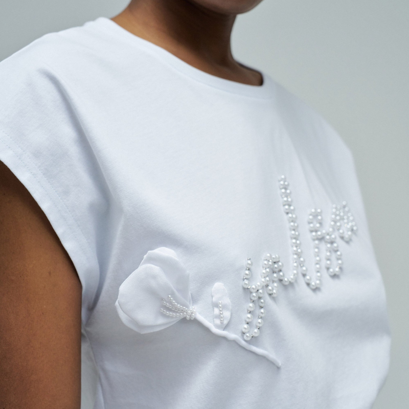 T-shirt branca de corte regular que combina elegância e conforto. Apresenta decote redondo clássico e mangas cava, criando uma silhueta versátil e moderna. O destaque fica por conta do branding em pérolas e do delicado aplique de flor, detalhes que elevam o design e conferem um toque sofisticado à peça. Perfeita para looks do dia a dia ou ocasiões mais descontraídas, esta t-shirt é uma escolha versátil que se adapta a qualquer estilo pessoal.