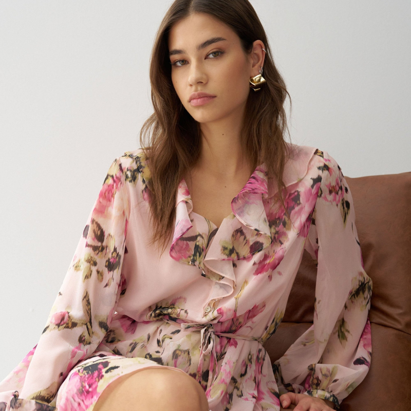 Vestido em tons rosa com padrão floral que combina elegância e movimento. O decote em V é realçado por folhos delicados, enquanto as mangas compridas apresentam punhos abotoados para um acabamento refinado. Destaca-se o cinto com pendentes nas extremidades, que adiciona dinamismo à silhueta. O folho na bainha confere volume e feminilidade. Vestido totalmente forrado para conforto e durabilidade. Uma peça versátil que transita com facilidade entre ocasiões casuais e mais sofisticadas.